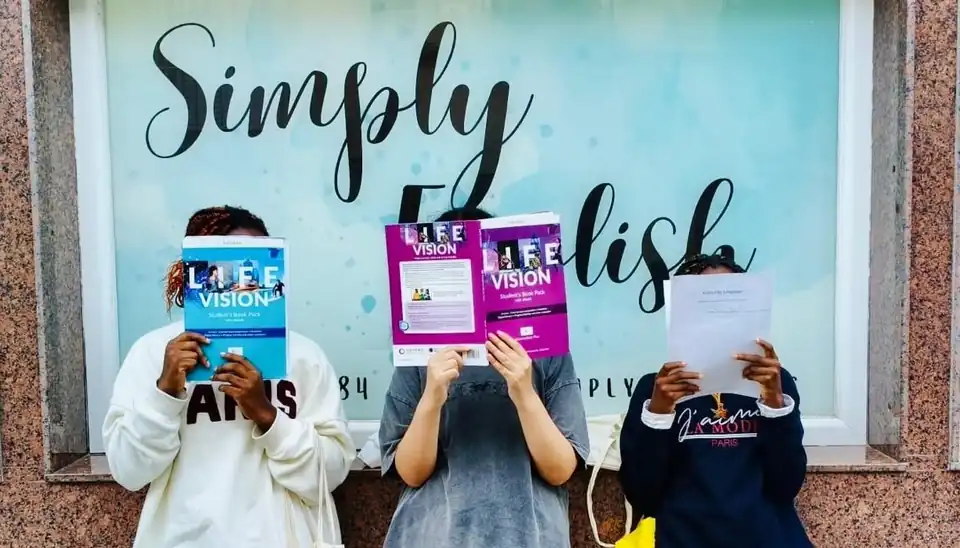 Tres alumnas delante del escaparate de Simply English, tapándose la cara con sus libros de inglés